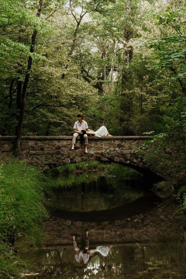 A couple on a stone arch bridge over a still creek in dense green woods. He sits barefoot with his legs over the edge, she lies back with her head in his lap