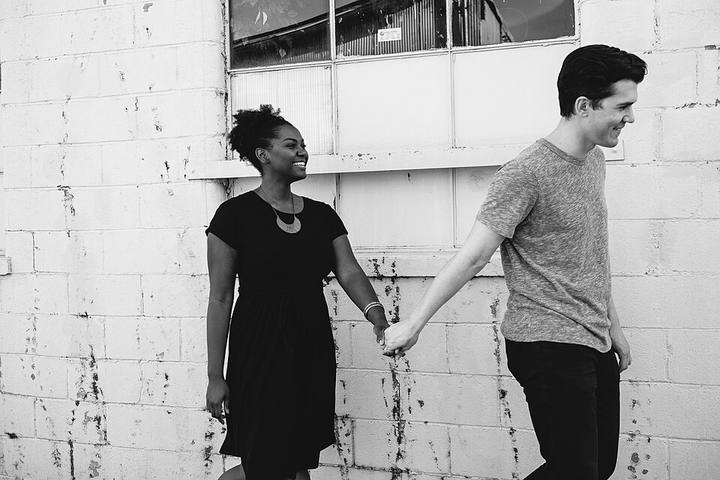 Black and white. A couple in front of a whitewashed brick wall, arms stretched between them still holding hands as they step apart, she is laughing