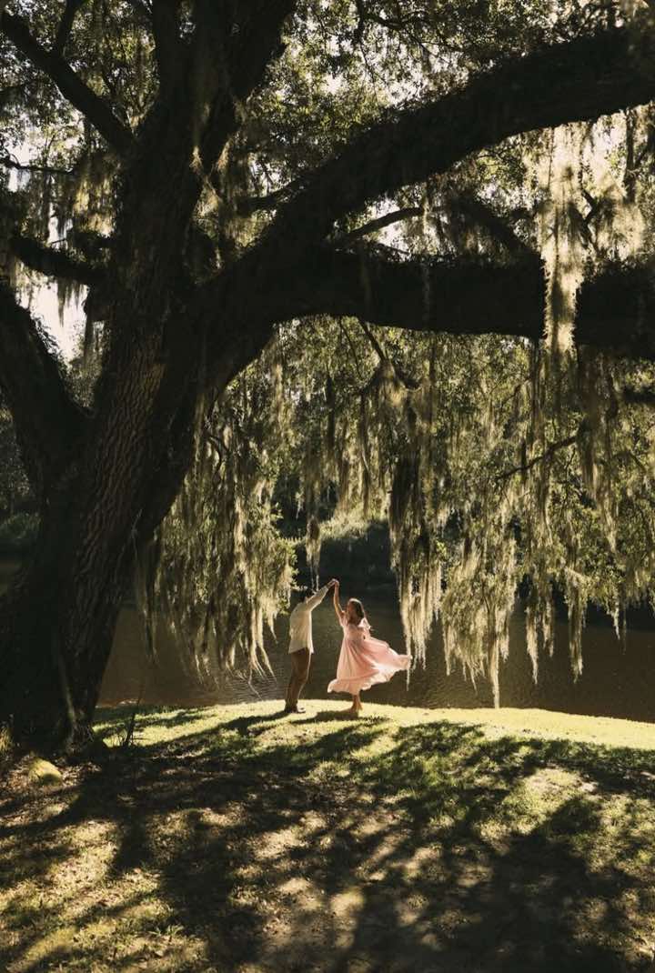 A couple on a lawn under a huge live oak hung with Spanish moss. He holds her hand up as she spins, her pale pink skirt flaring out, a lake behind them