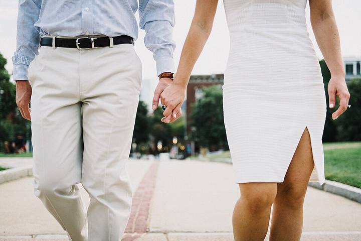 Cropped at the waist: a couple walking away down a paved path, hands joined between them, him in a pale blue shirt and cream trousers, her in a white dress